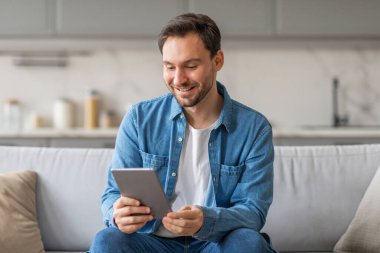 Mavi gömlekli ve kot pantolonlu bir adam oturma odasındaki açık gri koltukta oturur. Bir tablete bakıyor, gülümsüyor ve internette geziniyor ya da bir uygulama kullanıyor gibi görünüyor..