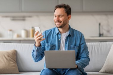 Beyaz bir kanepede oturan bir adam akıllı telefonuna bakarken gülümsüyor. O, mavi kot gömlek ve beyaz atlet giyiyor. Dizüstü bilgisayarı kucağında duruyor..