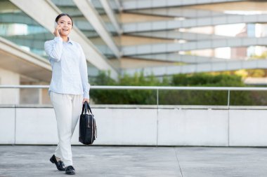İş kıyafeti giymiş profesyonel bir Asyalı kadın, akıllı telefonuyla konuşurken güvenli bir şekilde dışarıda yürüyor. Siyah bir çanta taşıyor, kopyalama alanı.