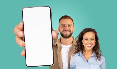Bir erkek ve bir kadın, deniz mavisi bir zemine karşı birlikte duruyorlar. Adam elinde kameraya doğru boş ekranlı bir akıllı telefon tutuyor. İkisi de gülümsüyor ve doğrudan kameraya bakıyorlar..