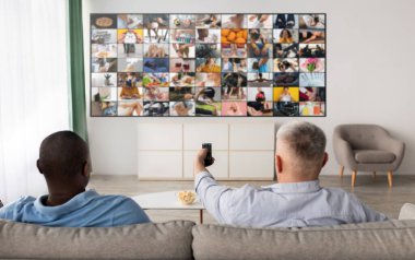 İki adam, biri siyah biri beyaz, oturma odasındaki bir kanepede oturuyor, büyük bir televizyon ekranına bakıyor. Ekran, mozaik etki yaratan bir görüntü ızgarası gösteriyor..