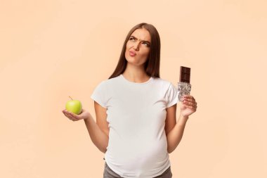 Hamilelik Beslenmesi. Yeşil Elma ile Beyaz Arkaplan 'ın üzerinde duran Çikolata arasında seçim yapan Bilmece Gibi Hamile Kız. Stüdyo Çekimi