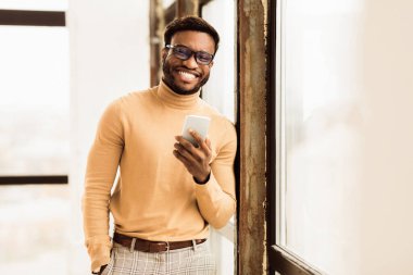 Kendine güvenen bir iş adamı. Kameraya gülümseyen, telefonu tutan, pencerenin yanında duran mutlu Afrikalı adam. Boşluğu kopyala