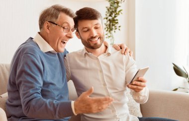 Orta yaşlı oğul, yaşlı babaya akıllı telefonu evde koltukta otururken nasıl kullanacağını gösteriyor. Seçici Odaklanma