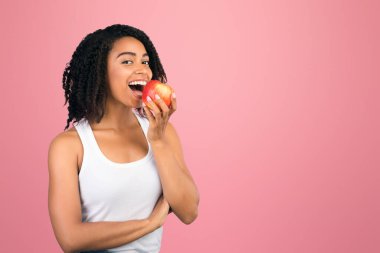 Diş sağlığı konsepti. Afrika kökenli Amerikalı bir kadın elma ısırıyor, meyve yiyor ve gülümsüyor. Boş boşluk