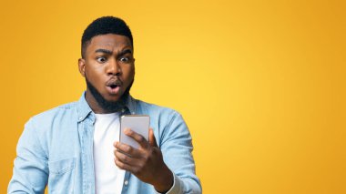 Beklenmedik bir mesaj. Şaşırmış Afro-Amerikan adam şok içinde akıllı telefon ekranına bakıyor, sarı arkaplanda fotokopi uzayıyla duruyor.