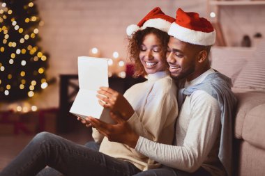 Noel hediyesini açan tatlı çift Noel ağacının önünde Noel Baba şapkası takıyor.