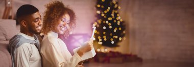 Xmas büyüsü. Neşeli siyah eşler Noel hediyesini birlikte açıyor, Noel ağacına karşı oturuyorlar.