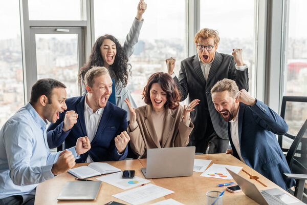 Разнообразная группа бизнес-профессионалов собирается вокруг ноутбука в современной офисной обстановке, все радуются и поднимают кулаки в честь праздника. Они в деловой одежде и кажутся взволнованными.