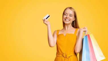 Genç alışverişkolik. Renkli alışveriş çantaları ve kredi kartı olan neşeli genç kızın portresi sarı arka planda, boş alanda izole edilmiş.
