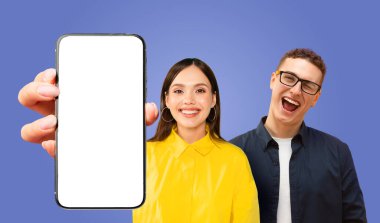Bir kadın kameraya gülümseyen beyaz ekranlı akıllı telefonu elinde tutuyor. Gözlüklü ve koyu renk gömlekli bir adam arkasında duruyor, gülümsüyor ve kameraya bakıyor..