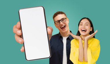 Bir erkek ve bir kadın, deniz mavisi bir arka planda yan yana durur. Adam sağ elinde akıllı bir telefon tutuyor, ekranı kameraya bakıyor. Kadın heyecanla gülümsüyor.
