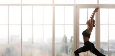 Evde yoga yapan atletik genç bir kadının profil fotoğrafı. Büyük bir panoramik pencere, boş bir yer.