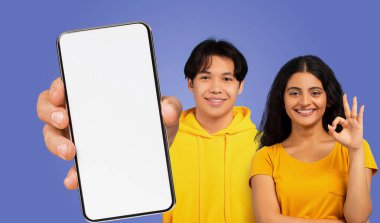 Genç bir Asyalı erkek ve Hintli kadın mavi bir arkaplanda yan yana duruyorlar. Kadın eliyle işaret verirken, adam elinde boş ekranlı büyük bir akıllı telefon tutuyor..