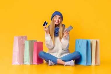 Güzel sarışın kız online alışveriş için akıllı telefon ve kredi kartı kullanıyor, alışveriş torbalarının arasında yerde oturuyor, pembe arka plan, fotokopi alanı