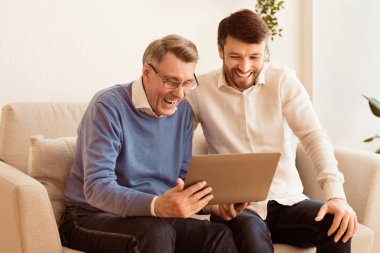 Orta yaşlı bir adam ve yaşlı bir baba dizüstü bilgisayar kullanıyor. Evdeki koltukta oturup gülüyorlar. Olgun Oğul ve Baba İlişkisi