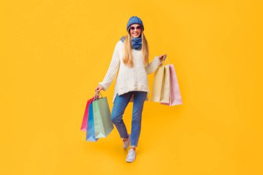 Kışlık elbiseli, güneş gözlüklü, pembe arka planda alışveriş torbaları taşıyan oldukça mutlu bir kız.