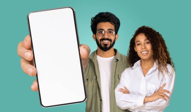 Hintli adam elinde beyaz ekranlı bir akıllı telefon tutuyor. Telefon ön planda ve arkasında bir adam ve kadın var. İkisi de kameraya gülümsüyor. Arkaplan sağlam bir deniz mavisi rengidir..