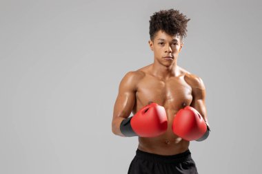 Genç bir atlet, kırmızı eldiven ve siyah şort giymiş, kendine güvenen bir boks duruşu içinde duruyor. Arka plan tarafsız, sporcuların odaklanması ve spordaki kararlılığı vurgulanıyor..