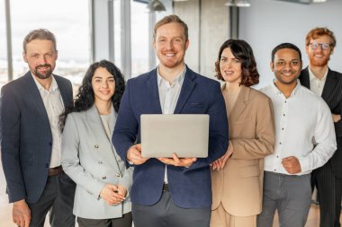 A diverse group of business professionals stand together in a modern office setting. The man in the center is holding a laptop and smiling at the camera, while the others smile and look at the camera clipart