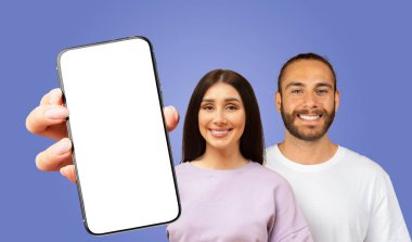 Genç bir kadın önünde boş bir ekranla akıllı telefonu tutuyor ve arkasında duran, gülümseyen genç bir adam. Arkaplan koyu mavi renkte..