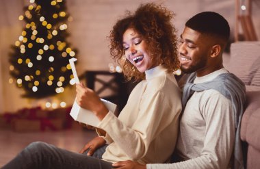 Heyecanlı Afro-Amerikalı çift Noel ağacının önünde oturma odasında Noel hediyesini açıyor.