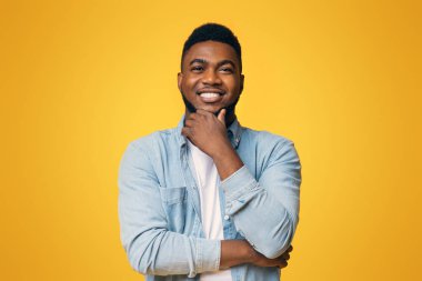 Portraif of handsome bearded african american man touching his chin and smiling over yellow studio background, copy space clipart