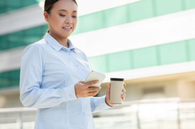 Asyalı kadın dışarıda açık mavi gömlekli bir elinde kahve fincanı diğer elinde akıllı bir telefonla dikiliyor. Modern bir şehir ortamında telefonunu kullanırken meşgul ve mutlu görünüyor..