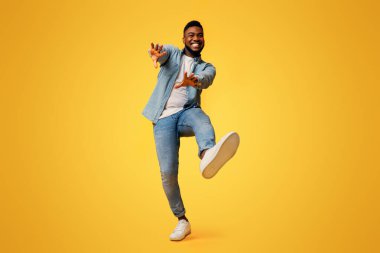 Komik Afrikalı Amerikalı adam dans ediyor ve ellerini sarı stüdyo arka planında kameraya uzatıyor.