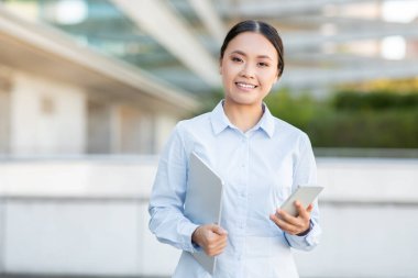 Genç, profesyonel bir Asyalı kadın şehir ticaret bölgesinde, bir elinde dizüstü bilgisayar, diğer elinde akıllı telefon tutarken güvenle gülümsüyor.