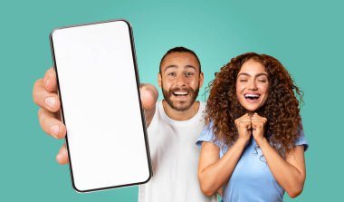 Genç bir çift turkuaz bir arka planın önünde duruyor. Adam elinde ekranı boş bir ekranla akıllı bir telefon tutuyor ve parmağını ekranı işaret ediyor..