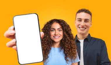 Uzun kıvırcık kahverengi saçlı genç bir kadın kameraya gülümseyen beyaz ekranlı bir akıllı telefon tutuyor. Kısa kahverengi saçlı genç bir adamın yanında duruyor.