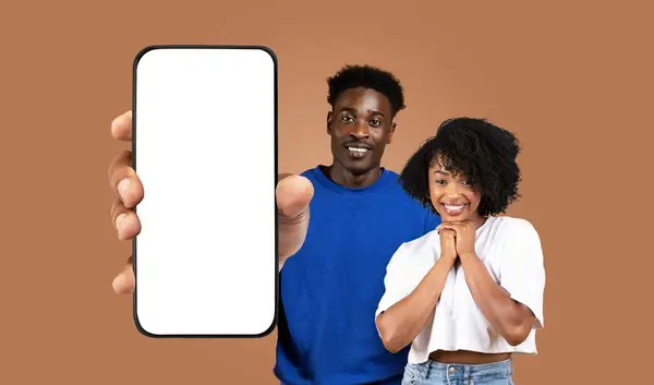 stock image African American young man and woman stand in front of a brown background, the man holding a smartphone with a blank screen in front of him. Both are smiling directly at the camera