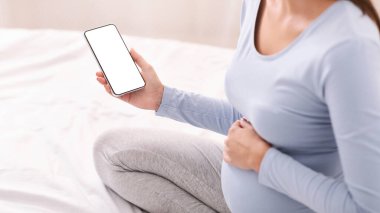 Hamilelik Uygulaması. Evde yatağında oturan boş ekranlı, cep telefonu tutan tanınmayan hamile bir kız. Kırpılmış, Mockup