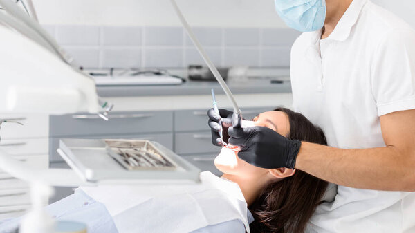 Young dentist doctor in glasses filling teeth to female patient, modern dental office, copy space