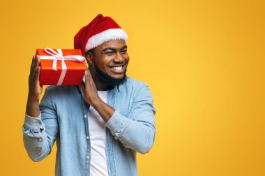 Tahmin et içinde ne var. Noel Baba şapkalı mutlu siyah adam Noel hediyesini sallıyor, hediye kutusundaki tıkırtıları dinliyor, sarı arka plan