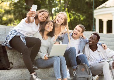 Şehirde dizüstü bilgisayarla çalışırken, merdivenlerde otururken akıllı telefondan selfie çeken mutlu uluslararası öğrenciler.