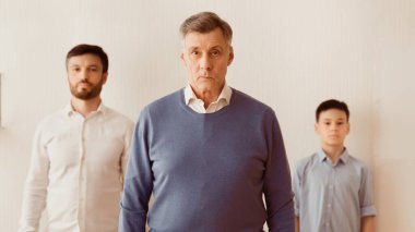 Büyükbaba, oğlu ve torunu arasında duruyor. Evdeki beyaz duvara bakan kameraya bakıyor. Panorama, Sığ Derinlik