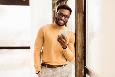 Genç Girişimci. Gülümseyen siyah iş adamı telefonu tutuyor, pencerenin kenarında duran kameraya bakıyor. Boş boşluk