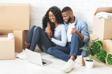 Taşınma ve taşınma konsepti. Neşeli Afrikalı çift yeni dairelerinde oturmuş dizüstü bilgisayarlarında film izliyorlar.