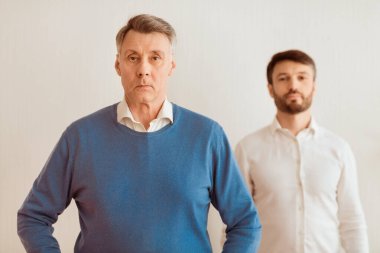 Otoriter ebeveyn. Yaşlı babası tarafından gölgede bırakılmış, beyaz arka plandaki kameraya bakan ciddi bir yetişkin oğul. Seçici Odaklanma