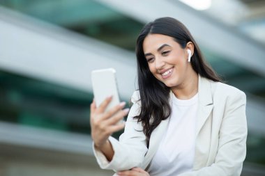 Uzun siyah saçlı ve beyaz ceketli bir kadın akıllı telefonunu kullanırken gülümsüyor. Beyaz kulaklık takıyor ve arka planda modern binalar olan bir şehir ortamında..