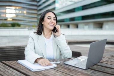 Beyaz ceketli ve beyaz tişörtlü genç bir kadın modern bir ofis binasının dışındaki ahşap bir masada oturuyor ve telefonuyla konuşurken gülümsüyor. Önünde bir laptop açık, kopyalama alanı