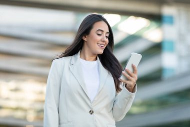 Beyaz ceketli ve beyaz gömlekli genç bir iş kadını şehir ortamında dikiliyor ve akıllı telefonuna bakıp gülümsüyor..