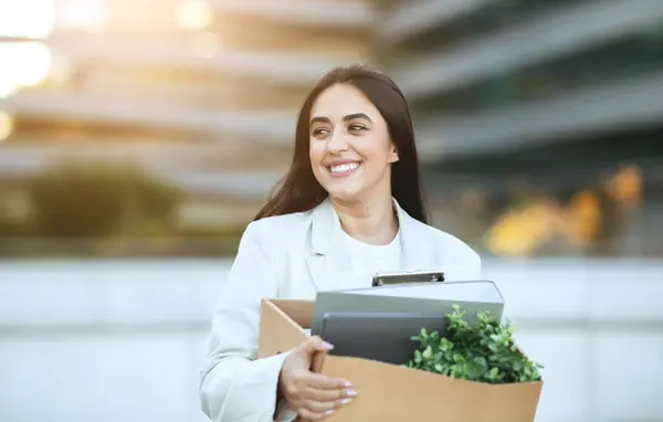 stock image A young woman in a white blazer smiles as she carries a box containing her belongings. She is leaving her office building and heading towards a new adventure, copy space