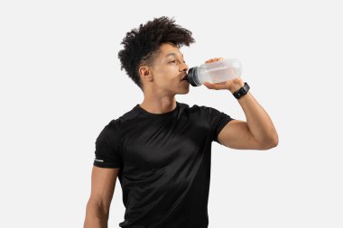 Genç bir adam susuz kalmadan antrenmanına ara veriyor. Siyah bir atletik gömlek giyiyor ve temiz bir su şişesinden su içmeye odaklanıyor. Arkaplan boş