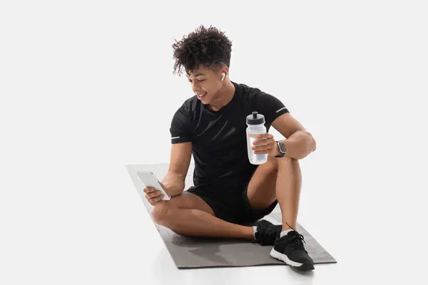 stock image A young adult, seated on a yoga mat, enjoys a moment of leisure by reading on phone while holding a water bottle. This guy demonstrates an active lifestyle, blending fitness with technology