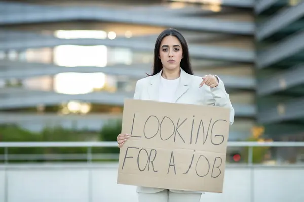 stock image A woman wearing a white blazer stands in front of a modern city building, holding a cardboard sign that reads Looking For A Job. She points at the sign with her right hand