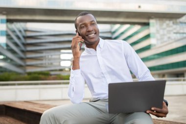 Afro-Amerikan iş adamı şehir ortamında ahşap bir bankta oturuyor ve aktif bir telefon görüşmesi yapıyor. Dizüstü bilgisayarı kucağına yerleştirilmiş.