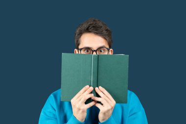 Genç bir adam koyu mavi arka planda oturuyor, önünde açık bir kitap tutuyor. Gözlük takıyor ve parlak mavi bir kazak giyiyor, kitabın tepesine bakıyor.
